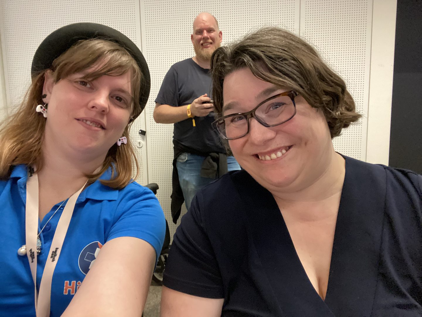 Foto van 3 mensen, 2 vrouwen in de voorgrond en 1 man in de achtergrond. De vrouw links, Marieke, heeft bruin schouderlengte haar, draagt een licht blauw shirt met een donker blauw en oranje logo van HiPerks erop, gamecontroller oorbellen en een zwart hoedje. De vrouw rechts, Marianne Dijkshoorn, heeft kort bruin haar, draagt een bril en heeft een zwart shirt aan. De man in de achtergrond, man van Marianne, is kaal met een baardje en draagt een zwart shirt met een blauwe jeans