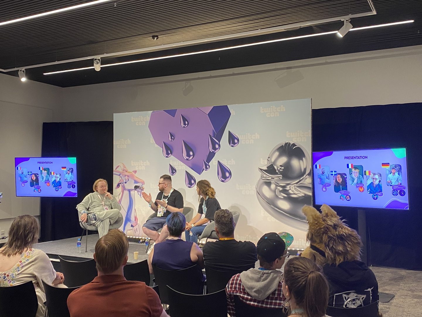 Foto van 3 mensen die in gebarentaal een panel houden over gebarentaal in streamen. aan beide kanten van het podium staat een scherm met foto's van 5 streamers die met gebarentaal streamen