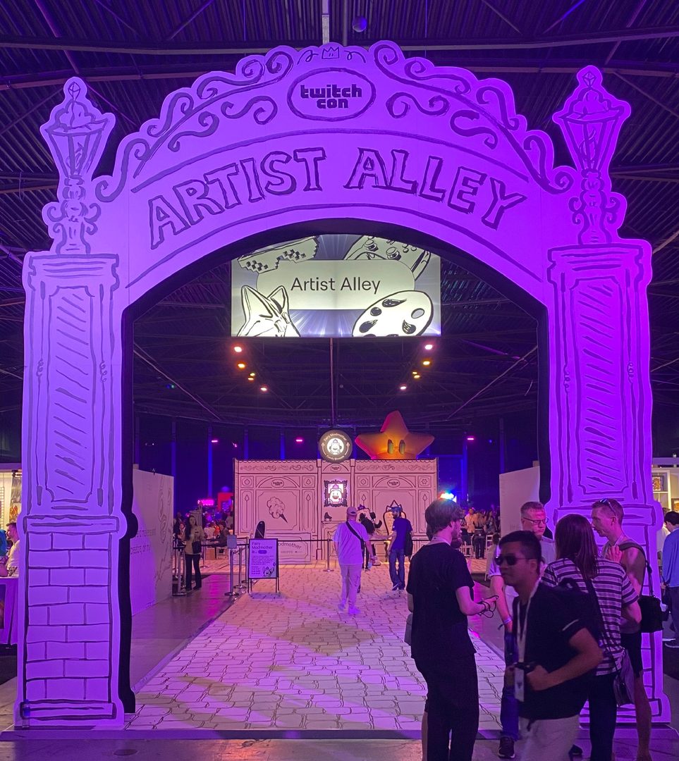 Foto van een poort uit karton waar de pilaren en boog op getekend zijn. De weg onder de boog lijkt ook getend net als de rest van de stand. Op de boog staan de woorden Artist Alley en Twitchcon