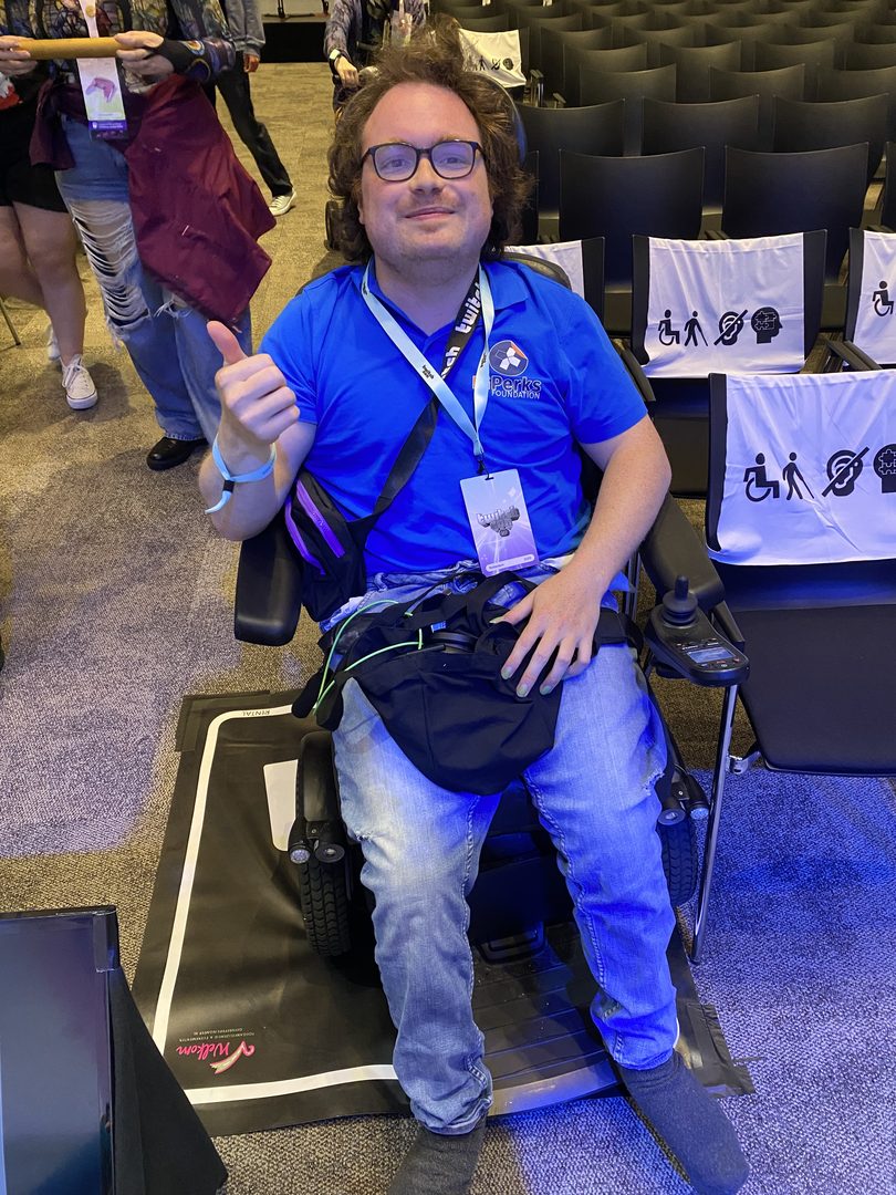 Foto van een man in een licht blauw shirt met donker blauw en oranje logo van HiPerks erop. Hij heeft bruin kort haar, draagt een bril en zit in een rolstoel die op een rolstoelmat staan